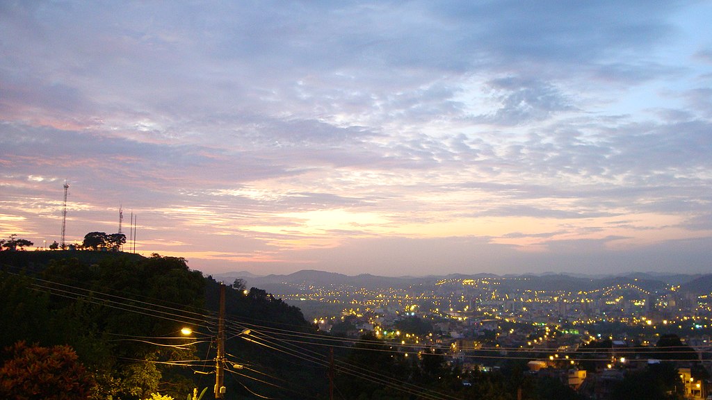 Montanha com a cidade iluminada ao fundo e o céu de fim de tarde azulado e um pouco avermelhado